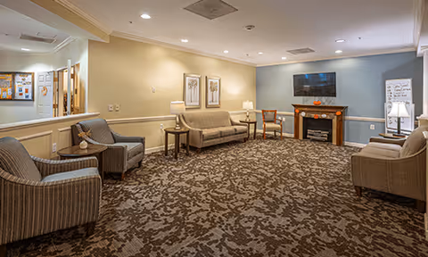 A cozy living room area with patterned carpet, two armchairs, a sofa, a wooden chair, and a fireplace with a TV mounted above it. The walls are painted in light beige and blue tones, and there are lamps and framed artwork on the walls. A whiteboard is visible near the fireplace.