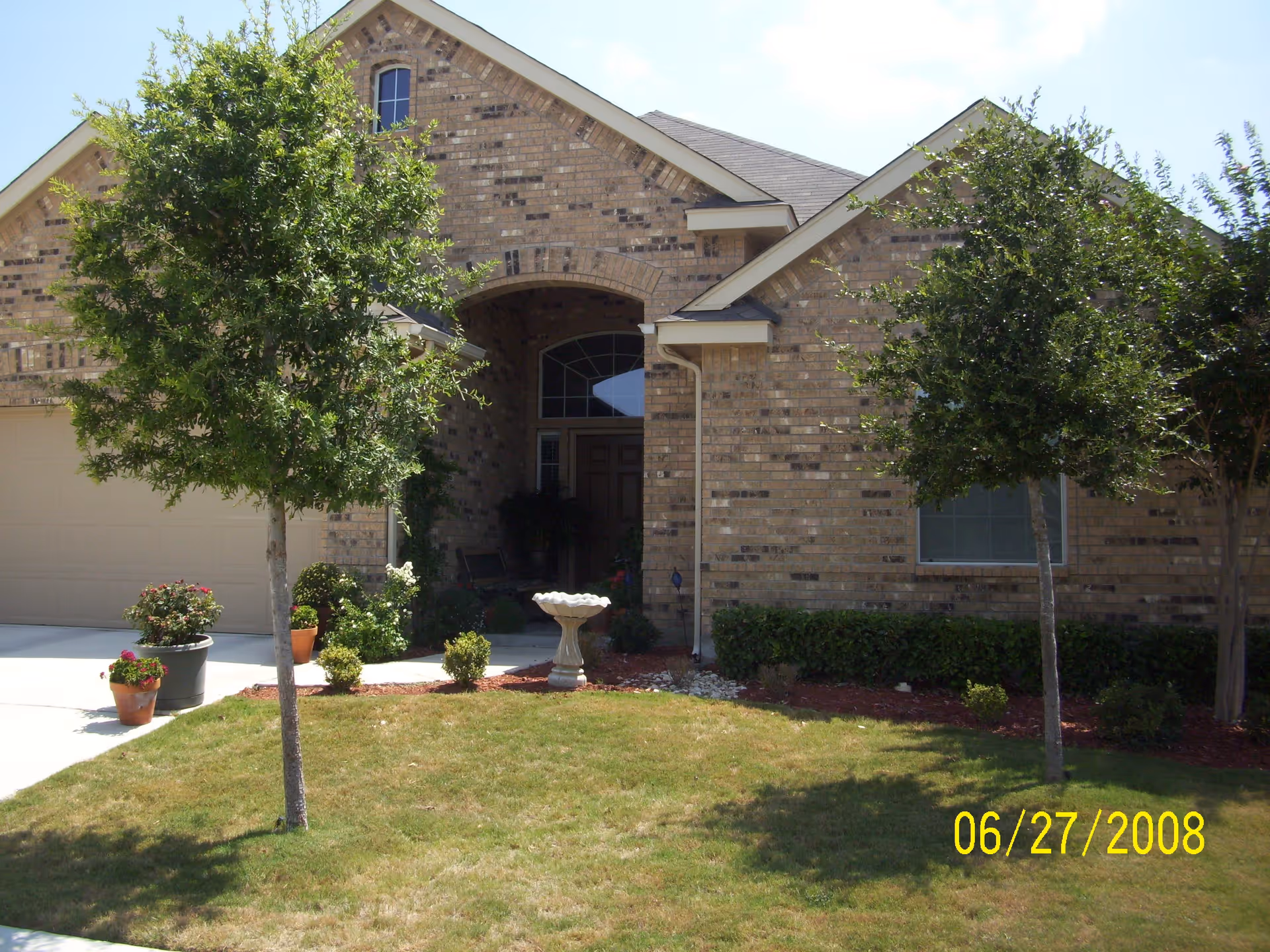 Front exterior view of a single-story brick house with a gabled roof, two small trees in the front yard, potted plants near the driveway, and a birdbath on the lawn. The entrance is recessed with an arched doorway and a brown door. The date 06/27/2008 is visible in yellow text at the bottom right corner.