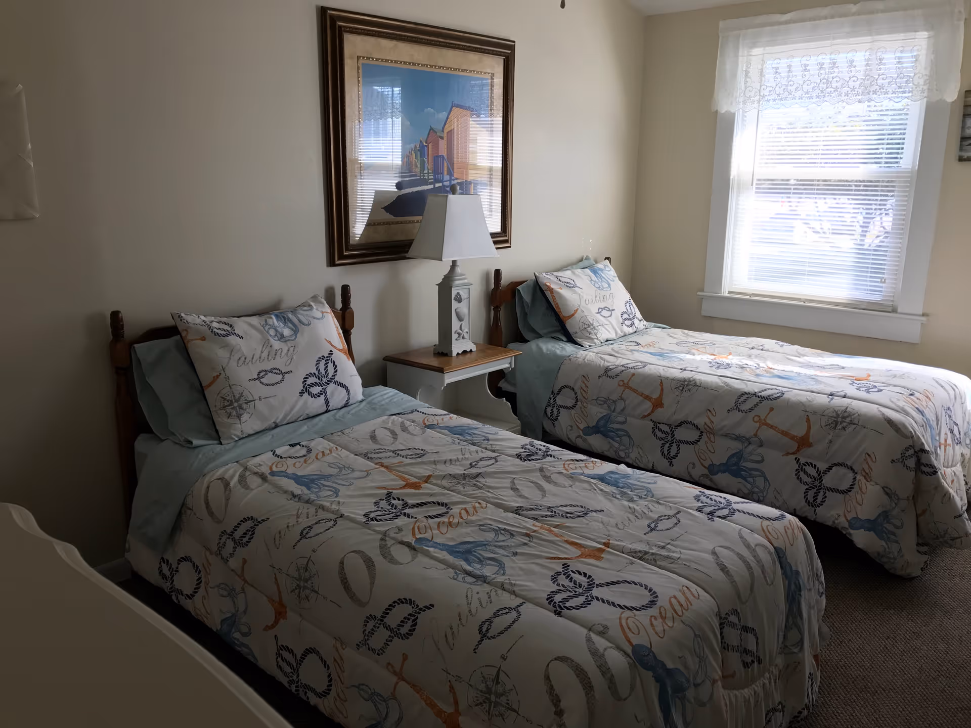 A bedroom with two twin beds featuring nautical-themed bedding with anchors, ropes, and octopus designs. A small wooden nightstand with a white lamp is placed between the beds. A framed picture hangs on the wall above the nightstand, and a window with blinds and a lace curtain is on the right side of the room.