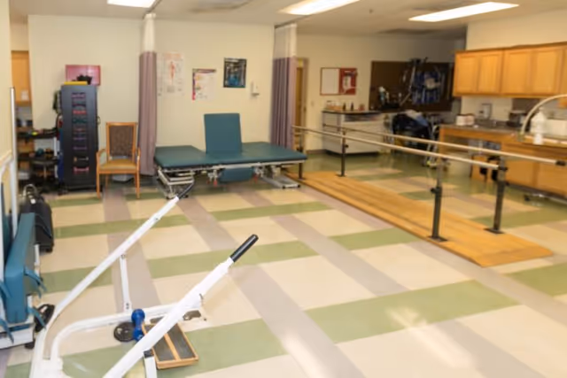 Physical therapy room with parallel bars for walking exercises, padded treatment tables, exercise equipment, chairs, and cabinets in a well-lit interior space.