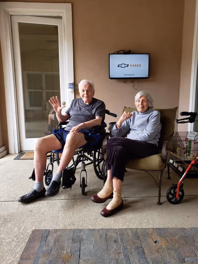 Two older adults seated on a covered patio — one in a wheelchair waving and the other in a chair next to a walker, with a small TV mounted on the wall behind them.