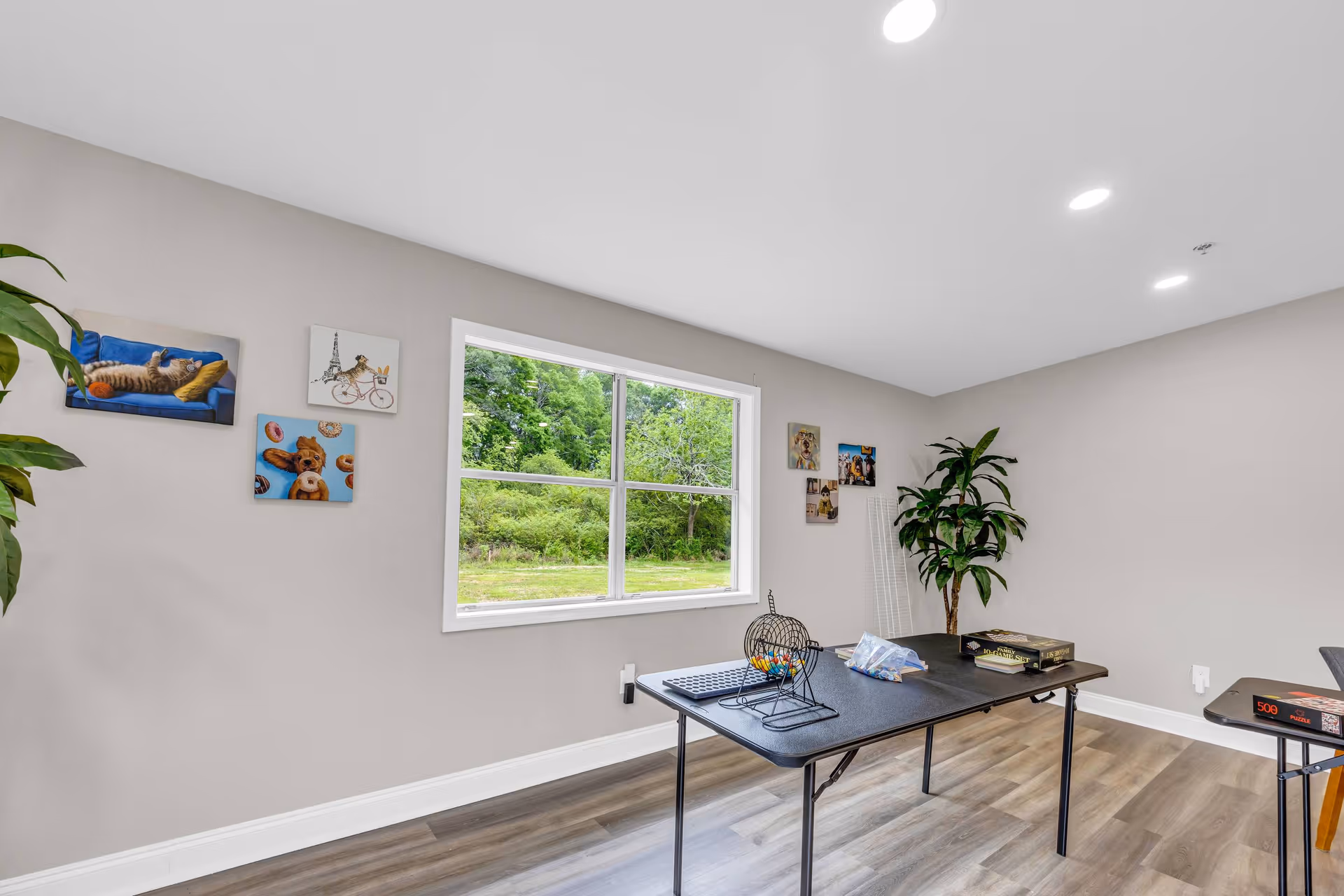 A bright room with light gray walls and wood flooring featuring a large window showing green trees outside. The room has two black folding tables with board games and game pieces on them. There are several playful animal-themed paintings on the walls and a tall green plant in the corner. The ceiling has recessed lighting.