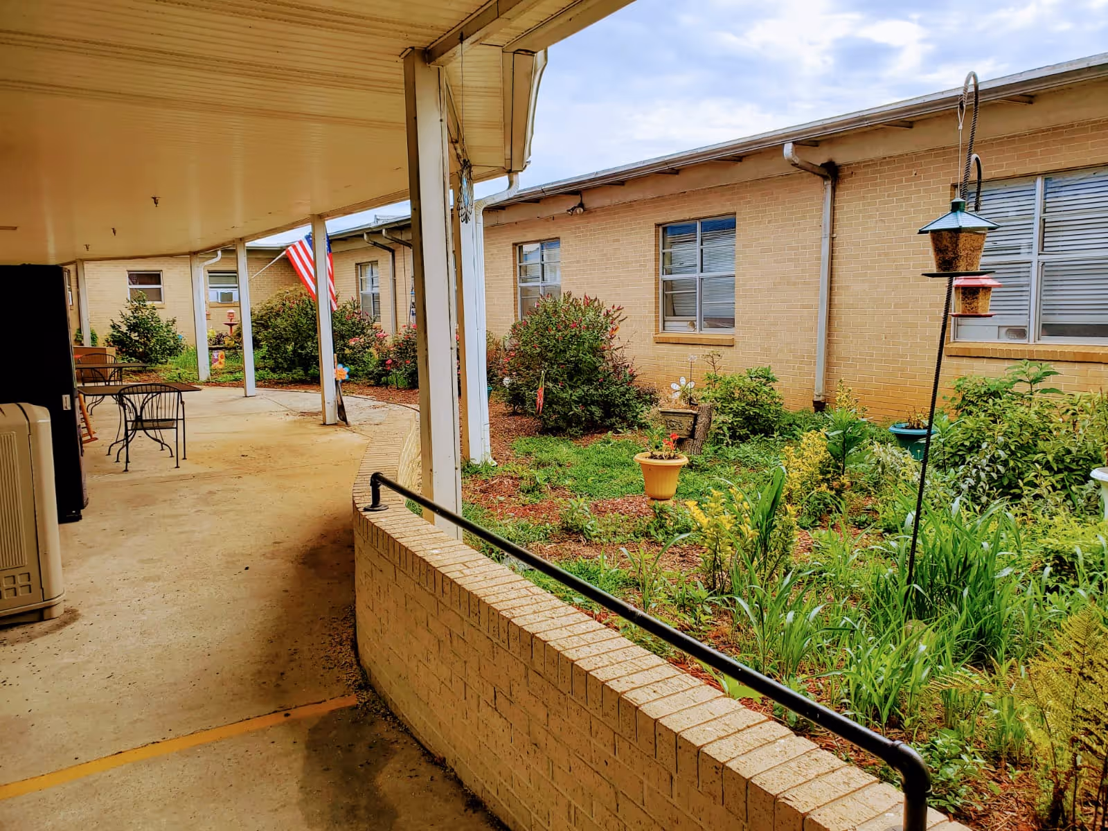 Covered courtyard walkway with patio tables, a planted garden bed, and a single-story brick building with windows.