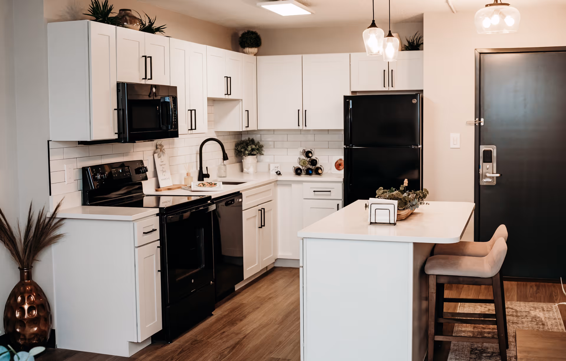 Modern kitchen with white cabinets, black appliances including a stove, microwave, dishwasher, and refrigerator. There is a white countertop island with two beige cushioned stools, pendant lights hanging above, and decorative plants on the counters and island. The floor is wood, and a dark-colored door is visible in the background.