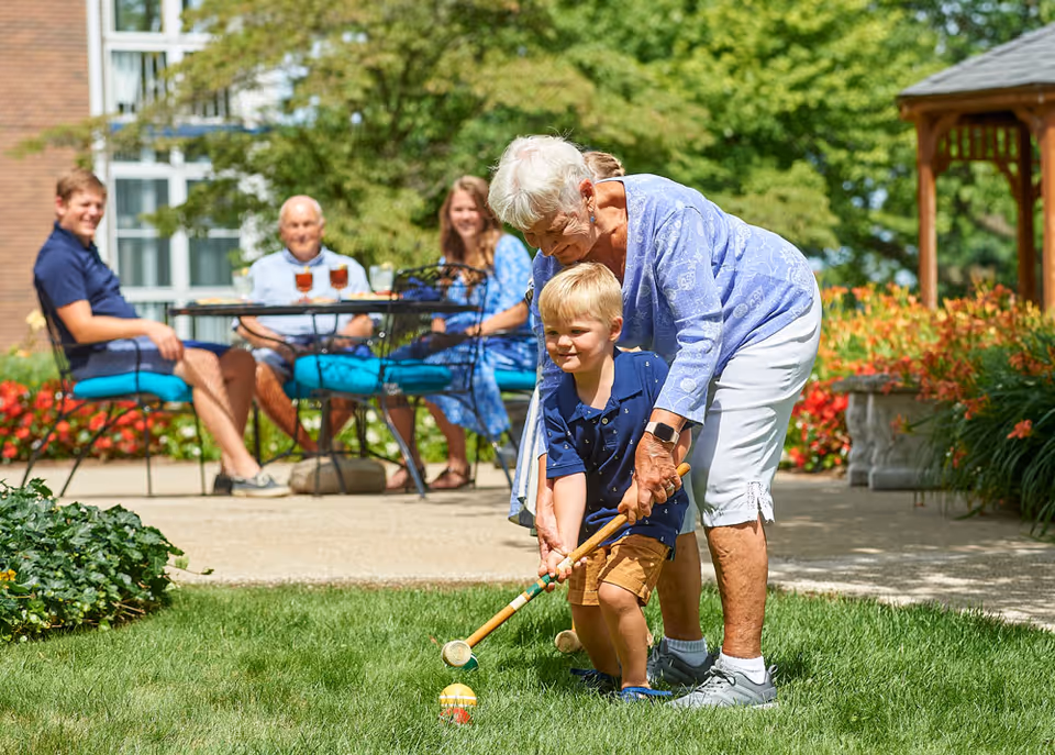 An elderly woman helps a young boy hold a croquet mallet on a grassy lawn. In the background, three adults sit at a table with drinks, smiling and watching the activity. The scene is set outdoors with greenery, flowers, and a wooden gazebo visible.