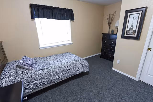 A simple bedroom with a single bed covered in a black and white patterned bedspread, a pillow, and a folded blanket. There is a window with a black valance letting in natural light. A black dresser with decorative items and a framed picture of the Eiffel Tower hang on the beige walls. The floor is carpeted in dark gray.