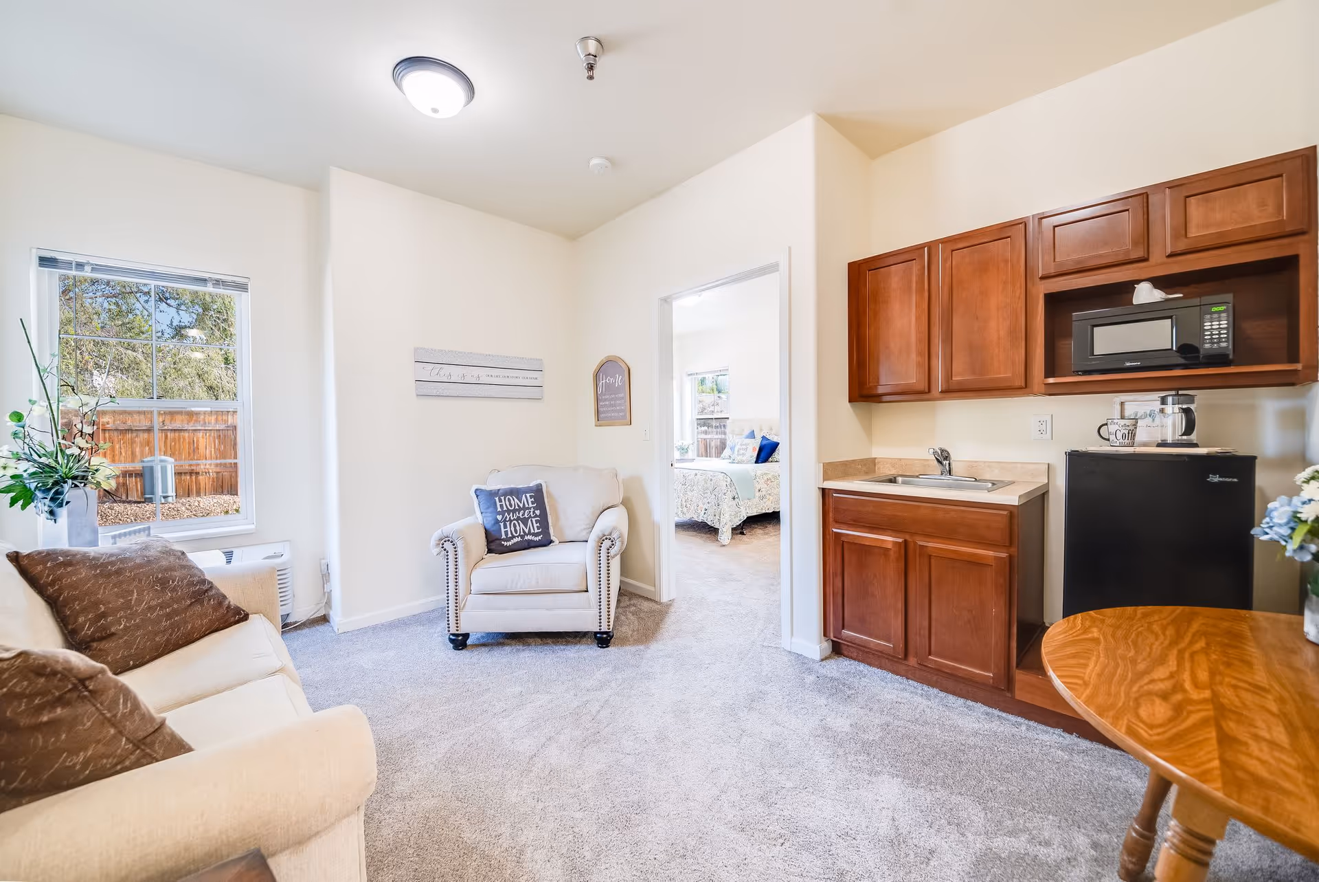 A bright and cozy assisted living apartment interior featuring a beige sofa with brown pillows, a beige armchair with a 'Home Sweet Home' pillow, a small kitchenette with wooden cabinets, a microwave, a mini fridge, and a sink. There is a window showing an outdoor view with a wooden fence and trees. A doorway leads to a bedroom with a bed and decorative pillows.