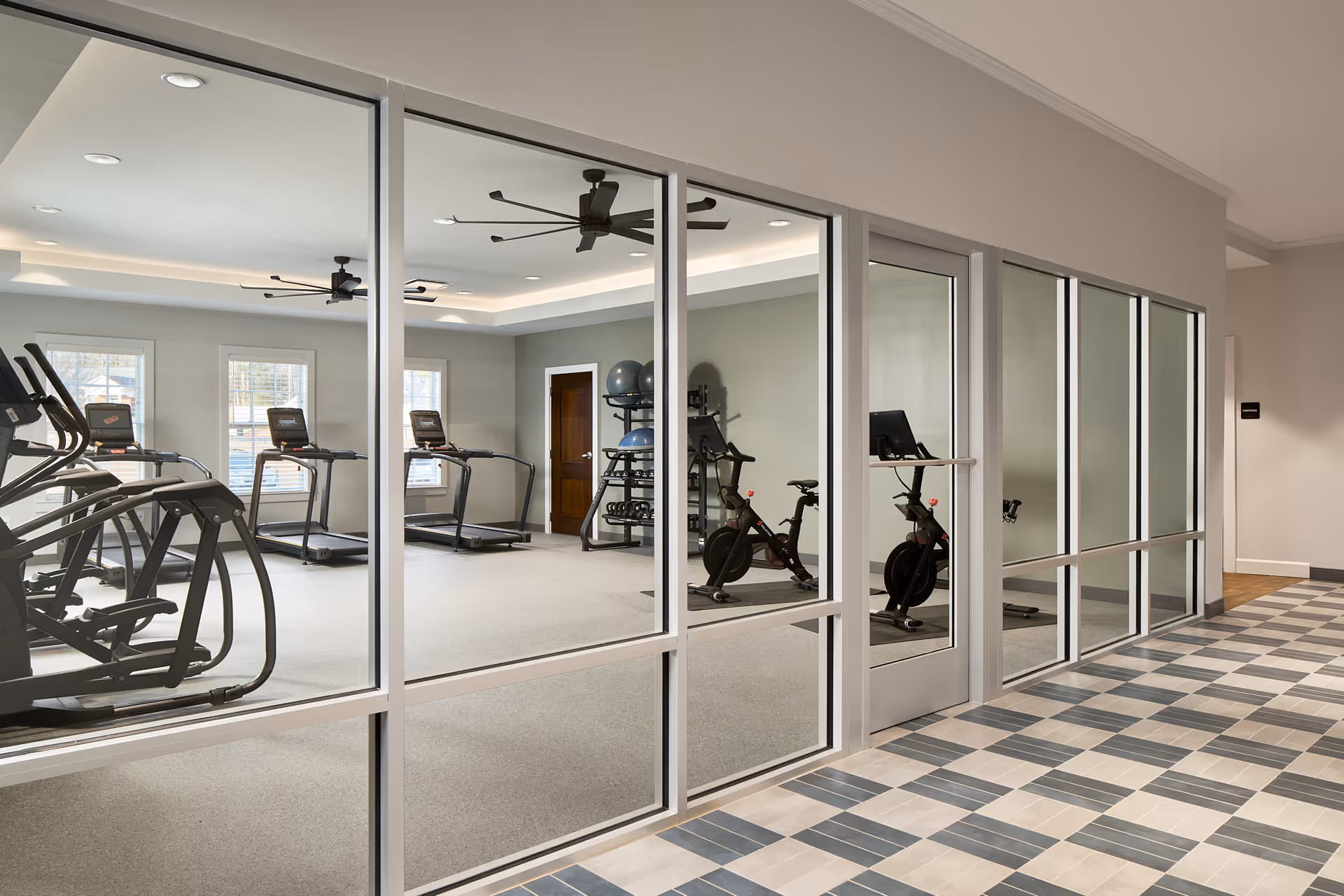 View through large glass windows into a fitness room with treadmills, stationary bikes, exercise balls, and weights. The room has ceiling fans and recessed lighting, with a checkered tile floor outside the fitness room.