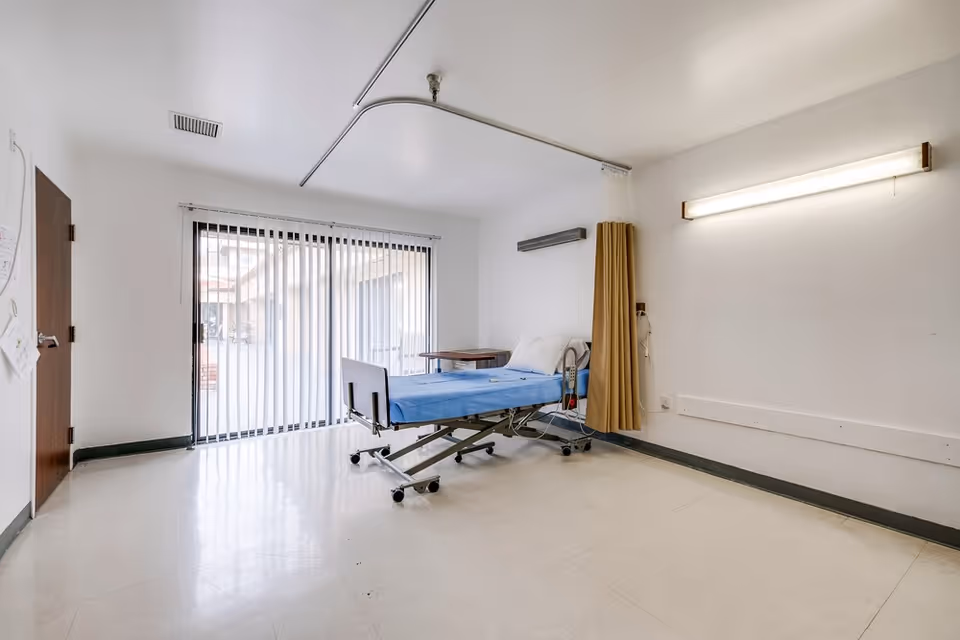 A simple hospital or healthcare facility room with a single adjustable bed covered in a blue sheet, a pillow, and a small wooden table next to it. The room has white walls, a large window with vertical blinds, a brown door, a fluorescent light fixture on the wall, and a privacy curtain partially drawn around the bed.