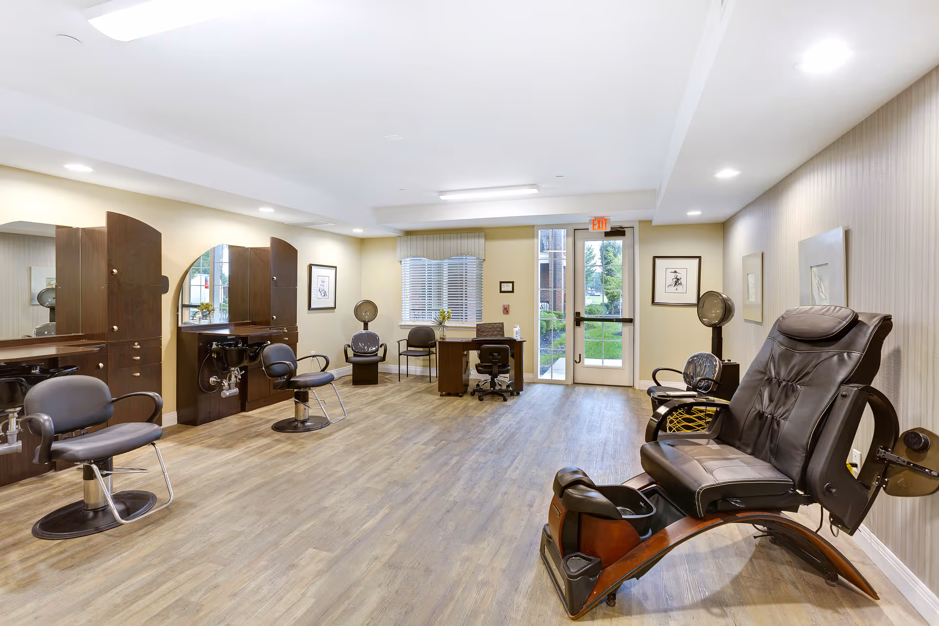 A spacious salon room with multiple styling chairs and mirrors along the left wall, a large black pedicure chair on the right, and a desk with chairs near a window and glass door at the back. The room has wood flooring, neutral-colored walls, and recessed lighting.