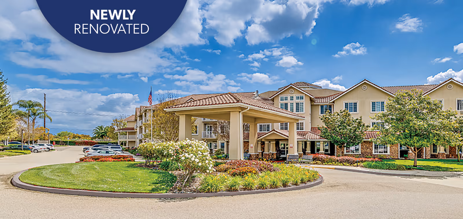 Front exterior of a newly renovated senior living building with a covered porte-cochère, circular driveway, and landscaped grounds under a blue sky.