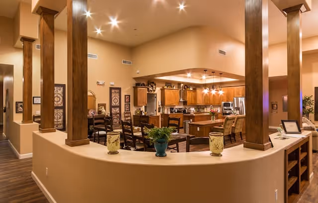 Interior view of a senior living facility showing an open-concept kitchen and dining area with wooden furniture, decorative plants, and warm lighting. The space features wooden columns, a kitchen island with pendant lights, and a cozy, inviting atmosphere.