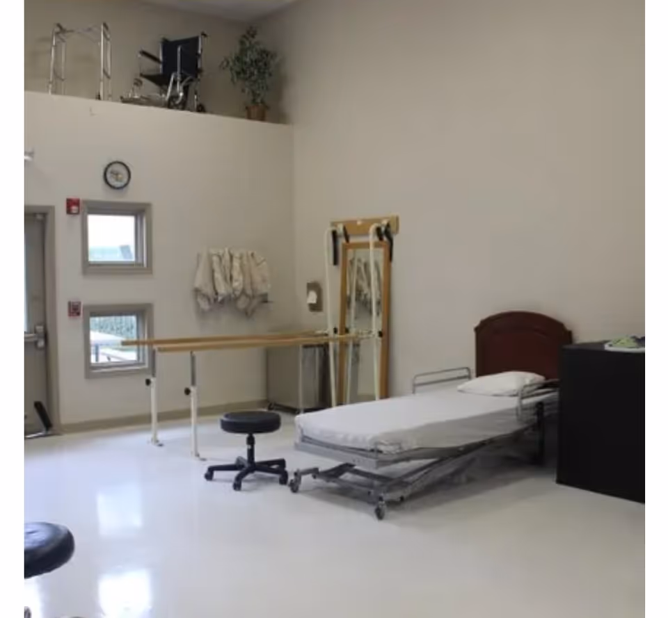A clinical room with a hospital bed, two black stools, parallel bars for physical therapy, a mirror with resistance bands, and a wheelchair and walker placed on a ledge near the ceiling. The room has white walls and a shiny white floor with two small windows and a clock on the wall.