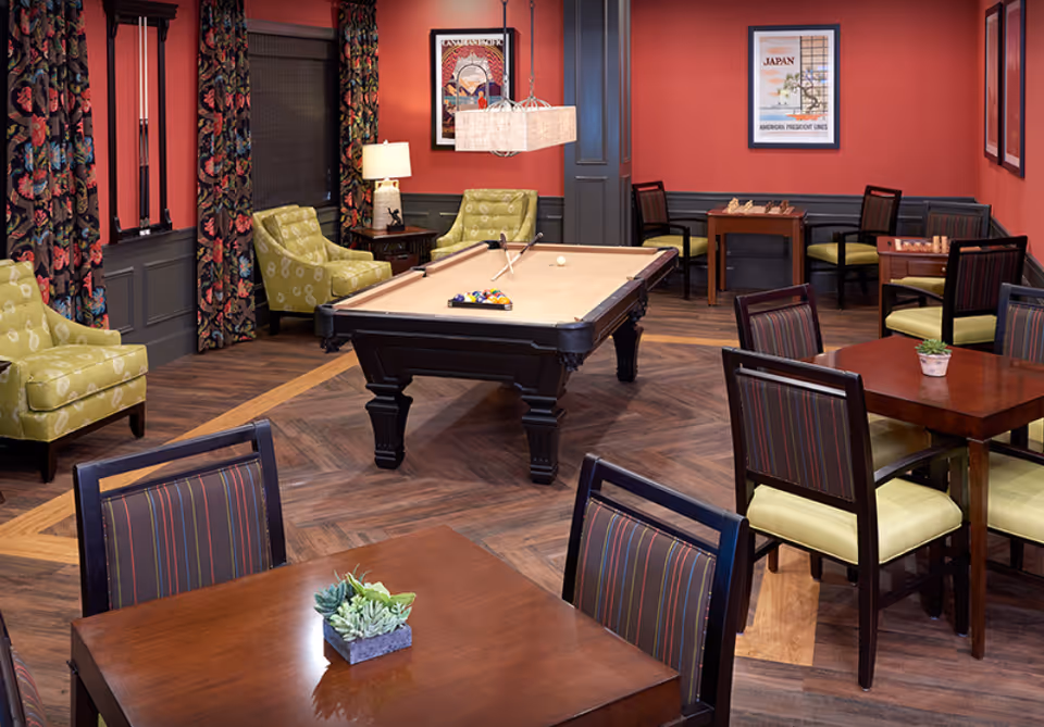 A cozy common room with a pool table at center, armchairs and several dining-style tables and chairs.