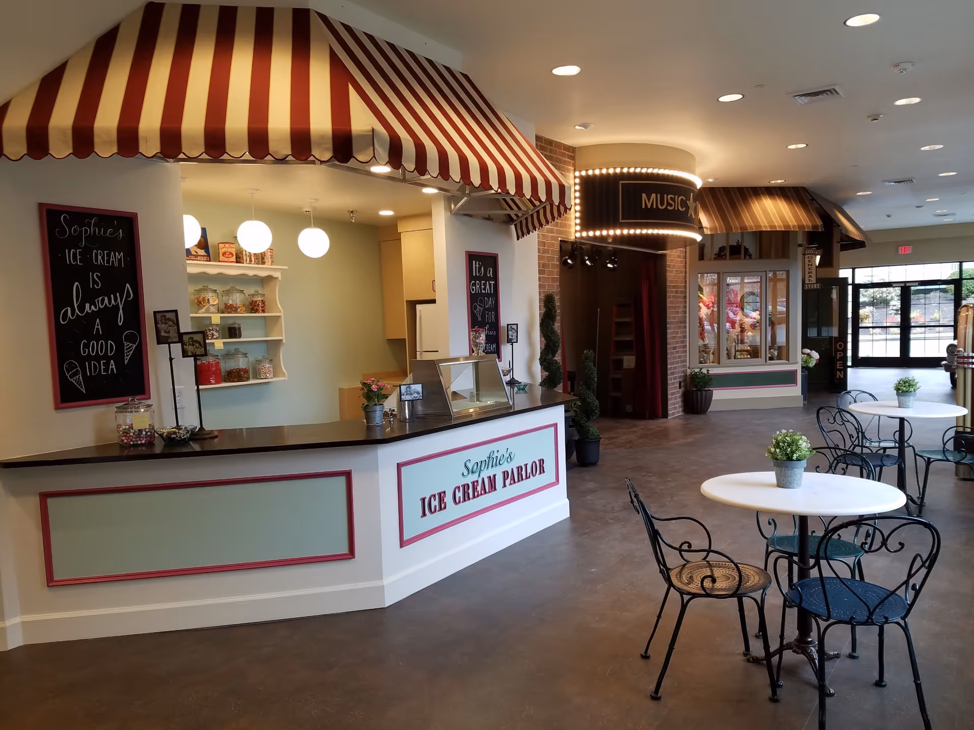 Interior view of a senior living facility featuring Sophie's Ice Cream Parlor with a striped red and white awning, a counter with candy jars, and a sign that says 'Sophie's Ice Cream is always a good idea.' There are round tables with wrought iron chairs and small potted plants. In the background, there is a section labeled 'Music' with marquee lights and another area resembling a general store with an 'Open' sign.