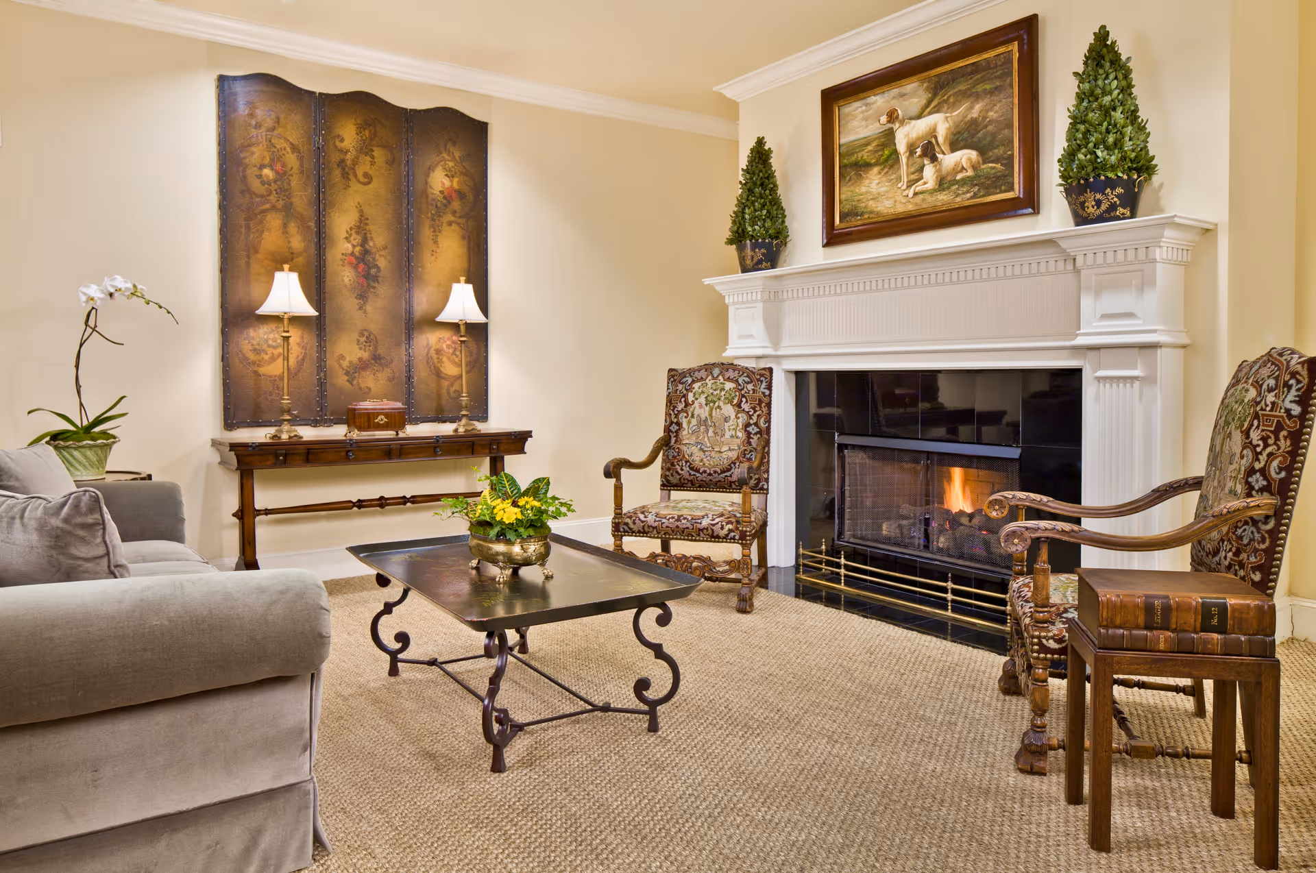 A cozy living room with a lit fireplace, two ornate wooden armchairs with patterned upholstery, a gray sofa, a metal coffee table with a plant centerpiece, a wooden console table with two lamps and decorative screen behind it, and a painting of two dogs above the fireplace mantel flanked by two potted plants.