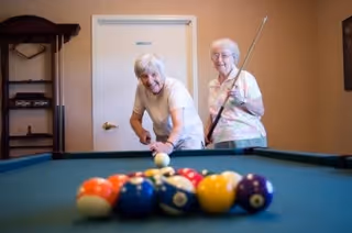 Two elderly women playing pool indoors, one is about to take a shot while the other watches and smiles.