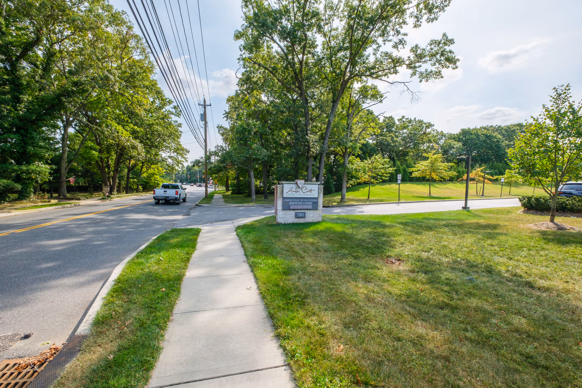 Sidewalk and road leading to the Amber Court entrance sign surrounded by trees and grassy grounds.