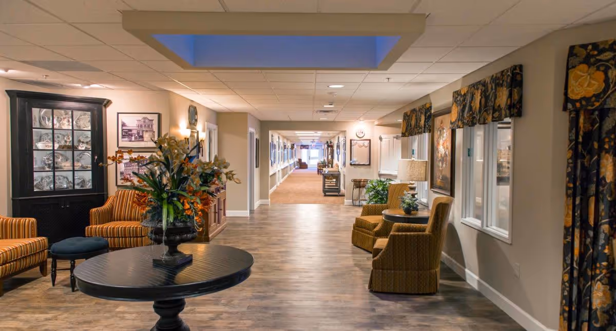 A well-lit hallway in a senior living facility with wooden flooring, comfortable seating areas including striped and patterned armchairs, a round table with a floral arrangement, framed pictures on the walls, and windows with floral valances.
