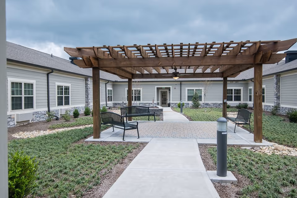 Outdoor courtyard area at Morning Pointe of Knoxville featuring a wooden pergola with benches underneath, surrounded by a paved walkway, grass, and landscaping. The building exterior has light gray siding with stone accents and multiple windows.