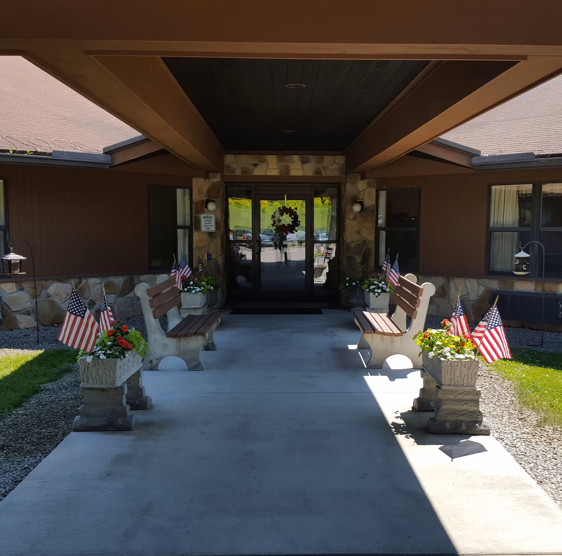 Covered entrance walkway to a building with two benches on either side, decorated with flower planters and small American flags. The building has stone and wood exterior walls, and glass double doors with a wreath hanging on them.