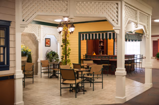 Indoor seating area with several tables and chairs arranged under a white lattice structure. There is a decorative streetlamp with hanging plants in the center. The walls are painted yellow with green trim, and there is a counter area with bar stools and striped green and white awnings. Potted plants and framed artwork decorate the space.