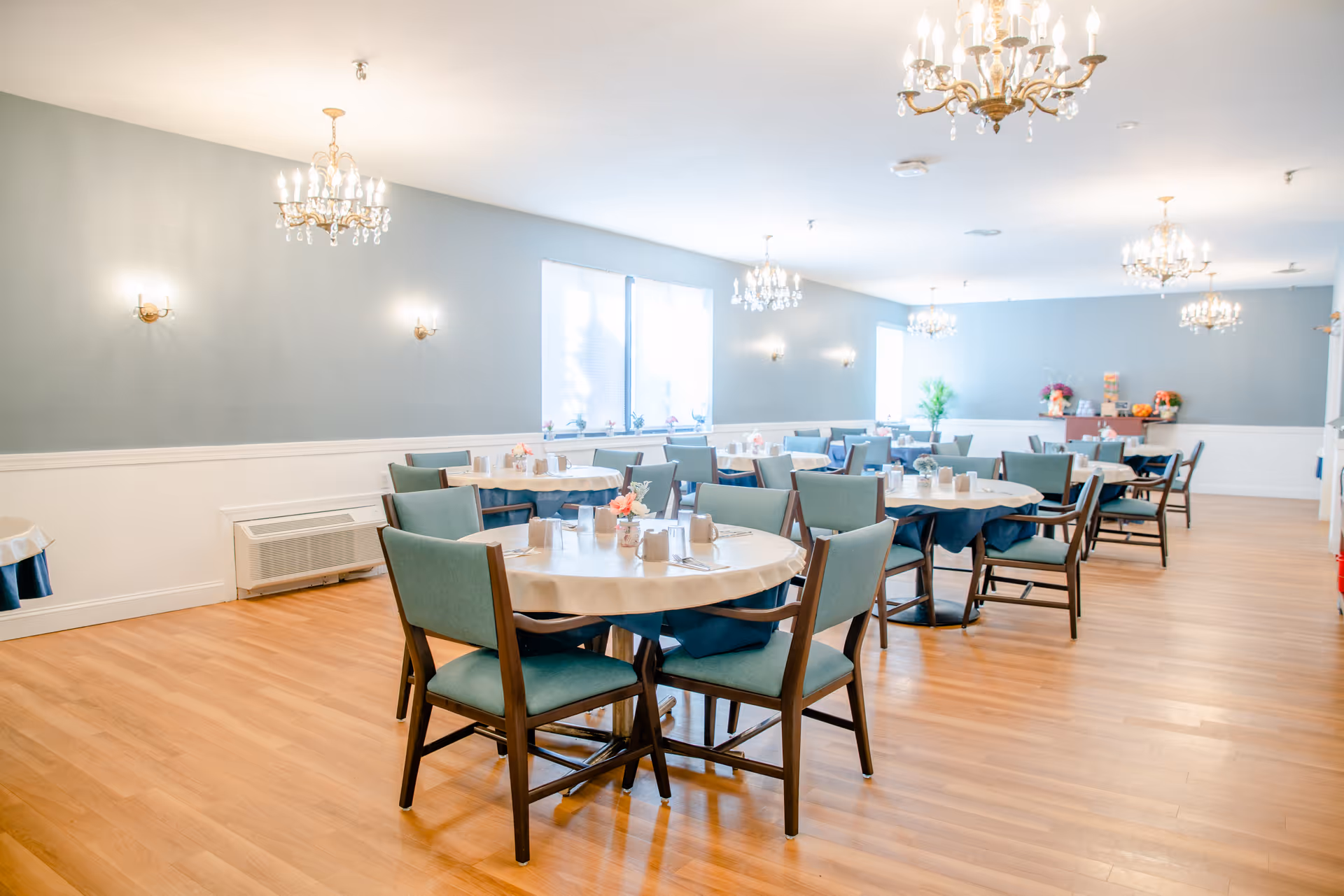 A bright and spacious dining room with multiple round tables covered with white tablecloths and blue underlays. Each table is set with cups, napkins, and small flower arrangements. The room features light blue walls with white wainscoting, wooden flooring, and several elegant chandeliers hanging from the ceiling. Large windows allow natural light to fill the space.