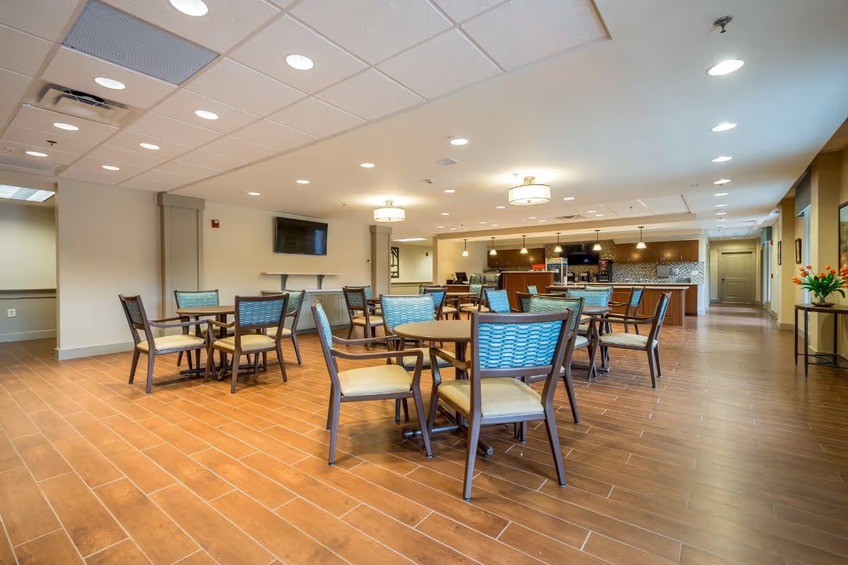 A spacious dining area with multiple round tables and chairs featuring blue patterned backs and beige seats. The room has wooden flooring, recessed lighting, and ceiling fixtures. In the background, there is a kitchen or serving area with pendant lights, a counter, and a tiled backsplash. A flat-screen TV is mounted on the wall to the left, and a hallway extends to the right.