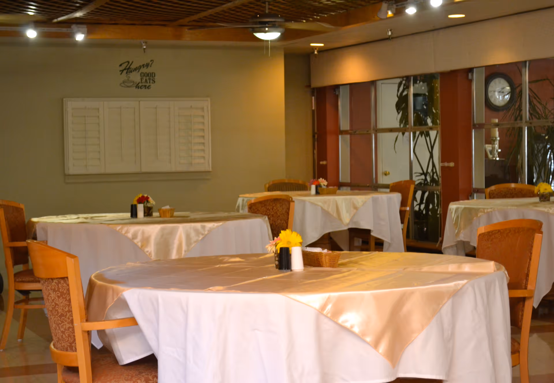 Interior view of a dining room with round tables covered in white and beige tablecloths. Each table has a small flower arrangement and condiments. Wooden chairs with patterned cushions surround the tables. The room has a wooden ceiling with recessed lighting and a wall clock is visible in the background.
