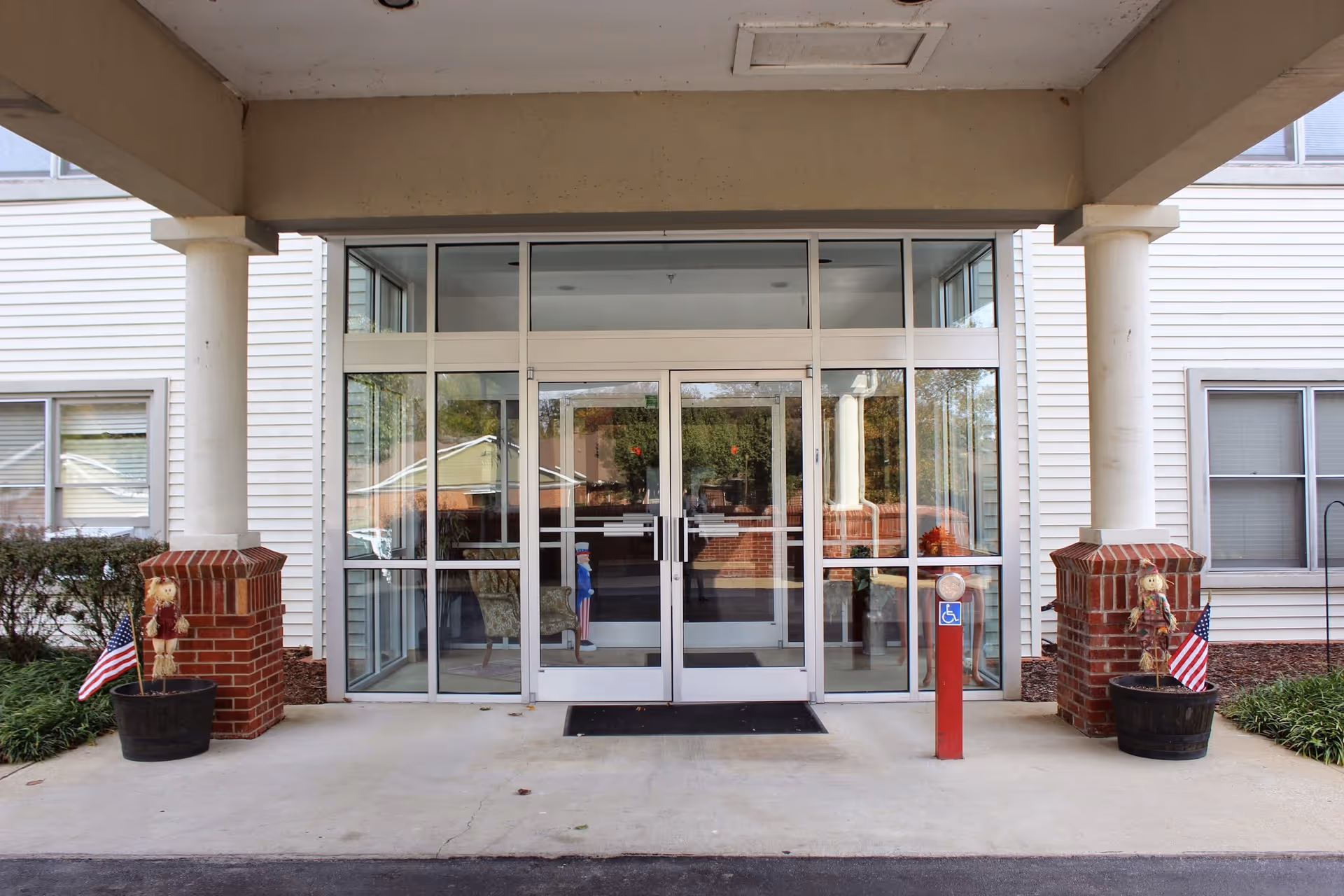 Covered main entrance to a senior living facility featuring glass double doors, brick pillars, and potted decorations with American flags.