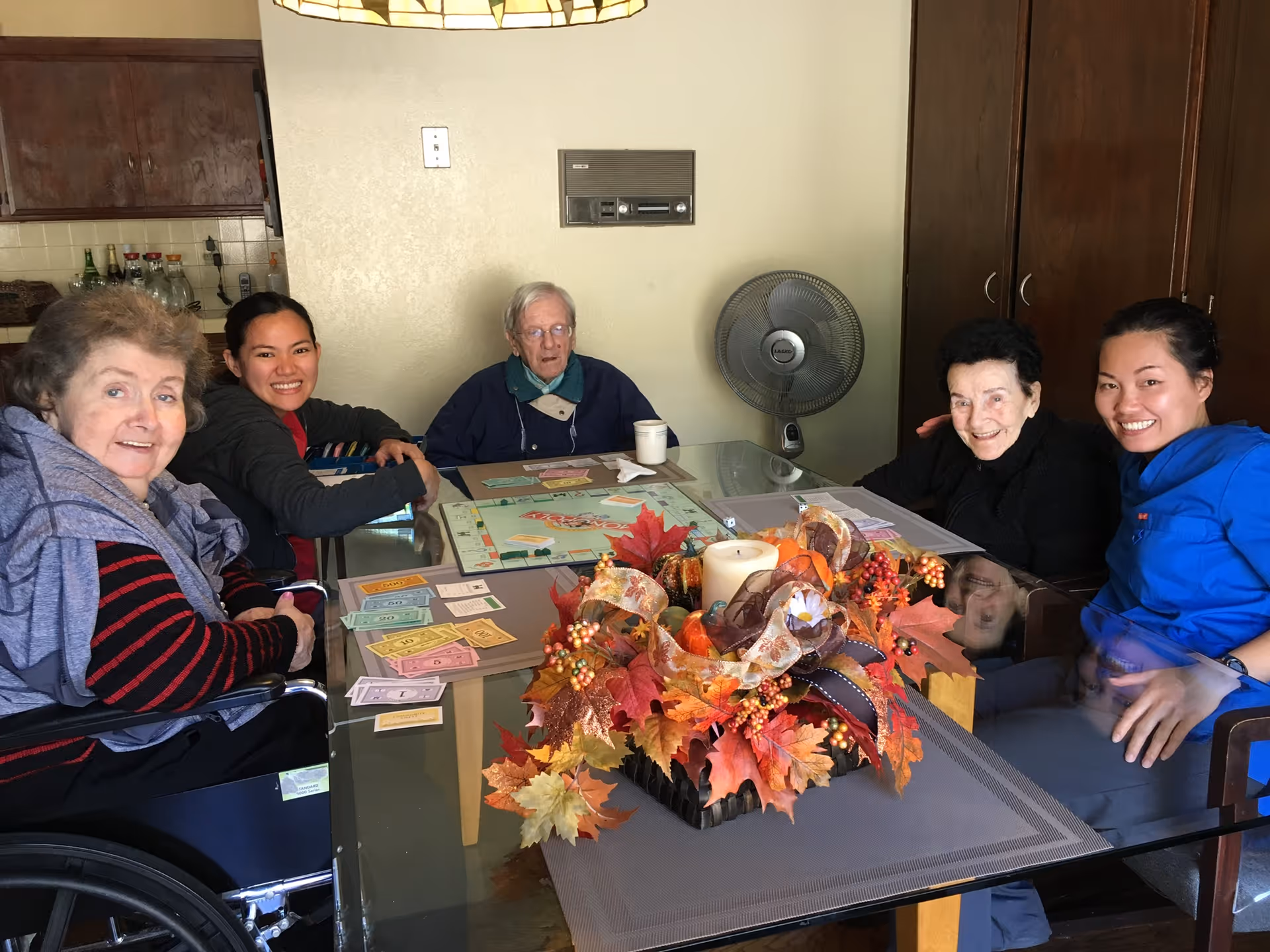 Two elderly women and one elderly man sitting around a glass dining table playing a board game with two younger women, in a room with wooden cabinets and a standing fan in the background. The table has a fall-themed centerpiece with leaves, berries, and a candle.
