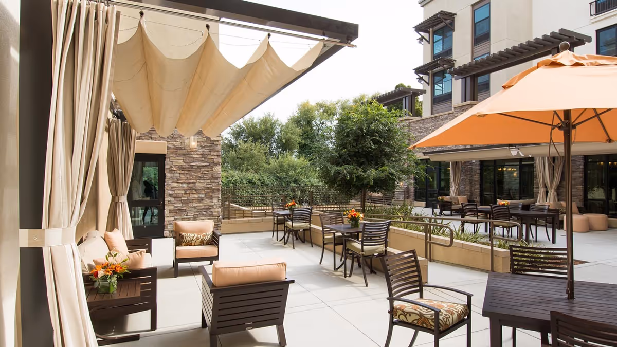 Outdoor patio area at Belmont Village Senior Living Albany featuring cushioned chairs, tables with flower arrangements, beige curtains, a large beige umbrella, and a stone building facade with greenery in the background.