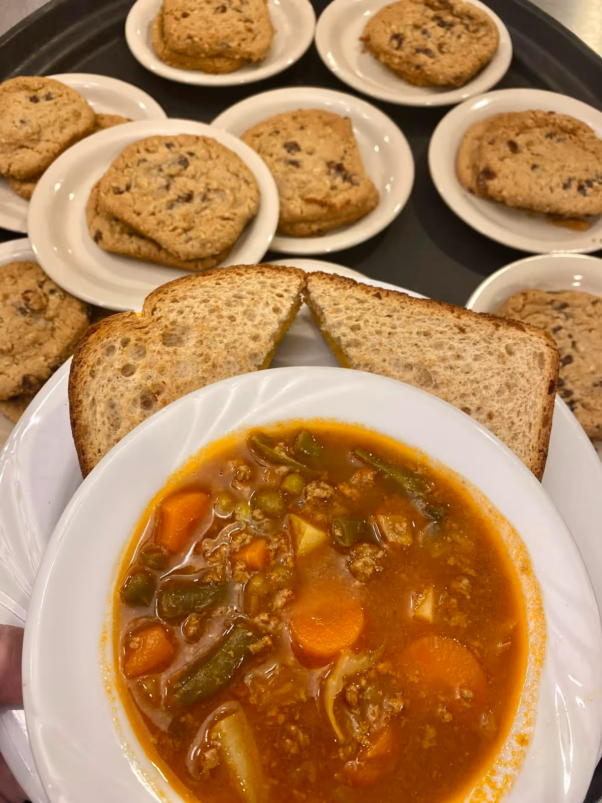 A white bowl filled with vegetable and beef soup, accompanied by two slices of toasted bread on the side. In the background, several small white plates each hold two chocolate chip cookies.