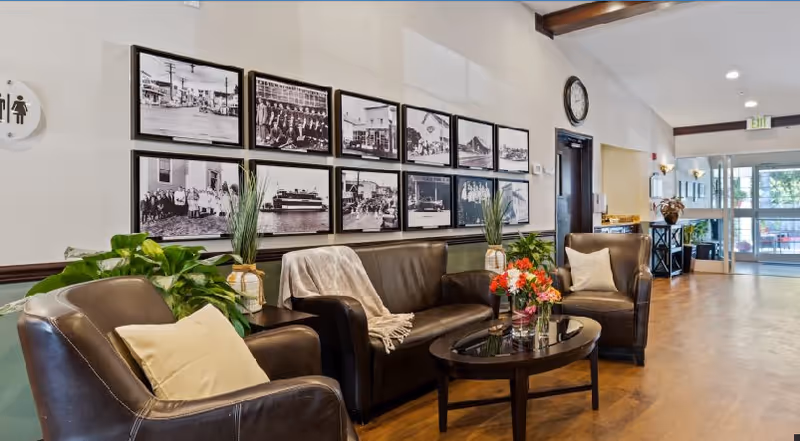 A cozy seating area in a senior living facility featuring two brown leather armchairs and a matching leather sofa with a beige throw blanket. A glass coffee table with a flower arrangement is in front of the sofa. The wall behind the sofa is decorated with a series of black and white framed photographs. There are green plants on either side of the sofa, and a clock is mounted on the wall above a door. The area is well-lit with natural light coming from glass doors in the background.