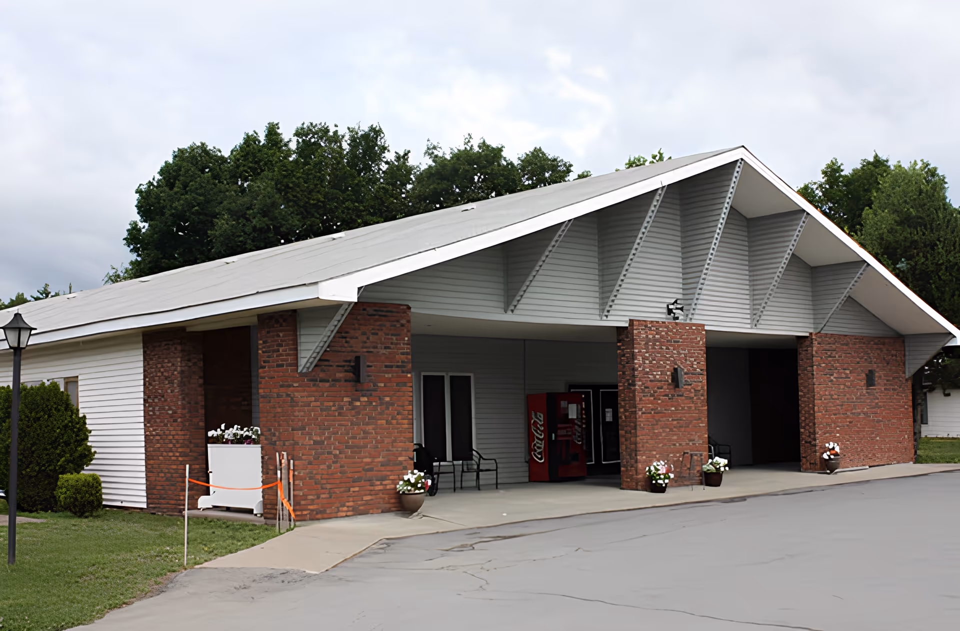 Exterior view of a building with a slanted roof, brick pillars, and white siding. There are potted flowers near the entrance, a Coca-Cola vending machine, and some chairs under the covered area. Trees and a cloudy sky are visible in the background.
