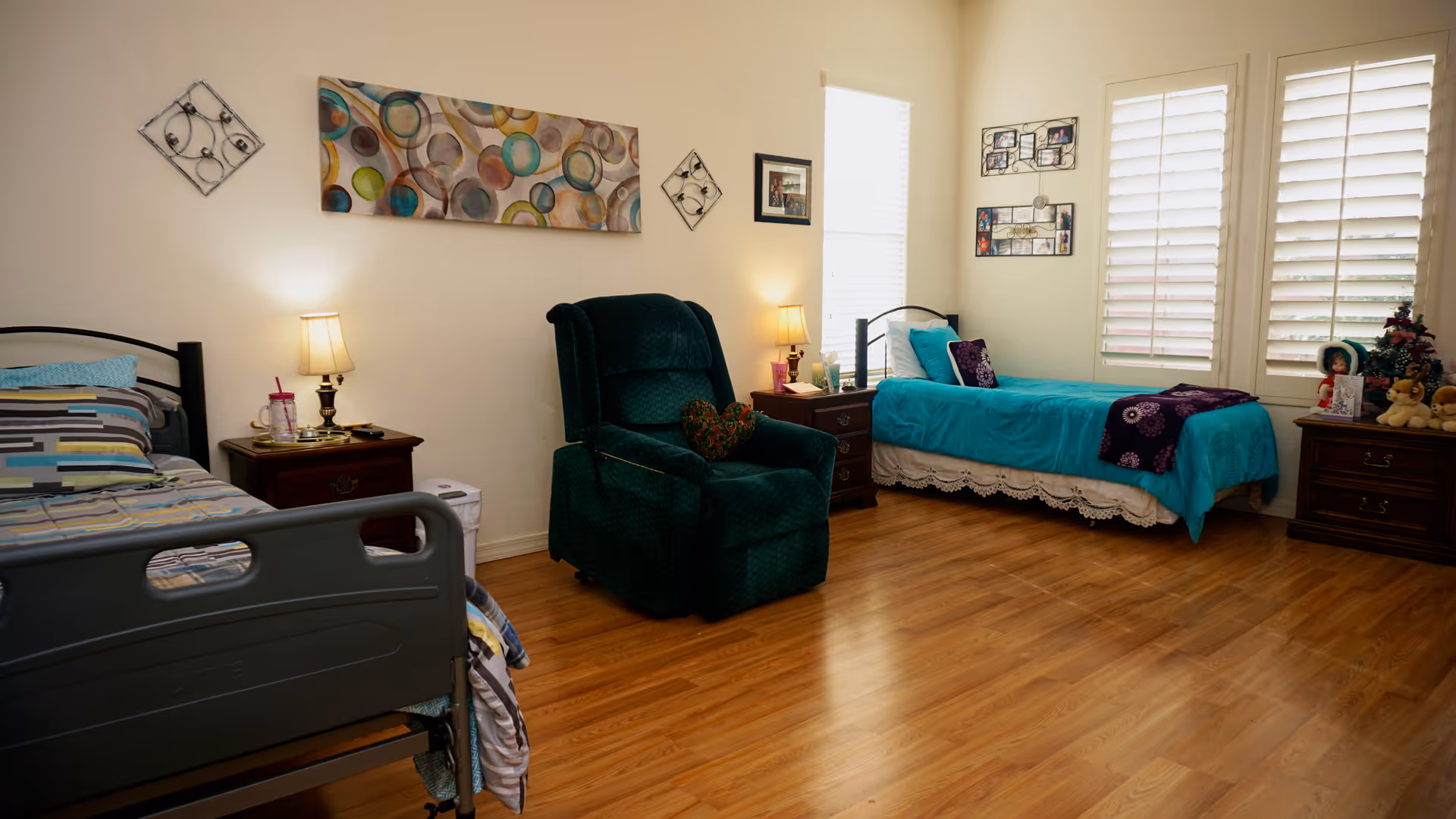 A well-lit bedroom with two beds, one with a striped bedspread and the other with a turquoise bedspread. Between the beds are two wooden nightstands with lamps and personal items. A dark green recliner chair is positioned in the center of the room. The walls are decorated with colorful artwork and photo frames. The floor is wooden, and there are windows with white blinds letting in natural light.