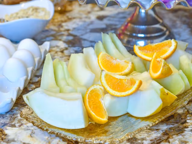 Sliced melon and orange wedges arranged on a decorative platter on a countertop with eggs in a tray nearby.