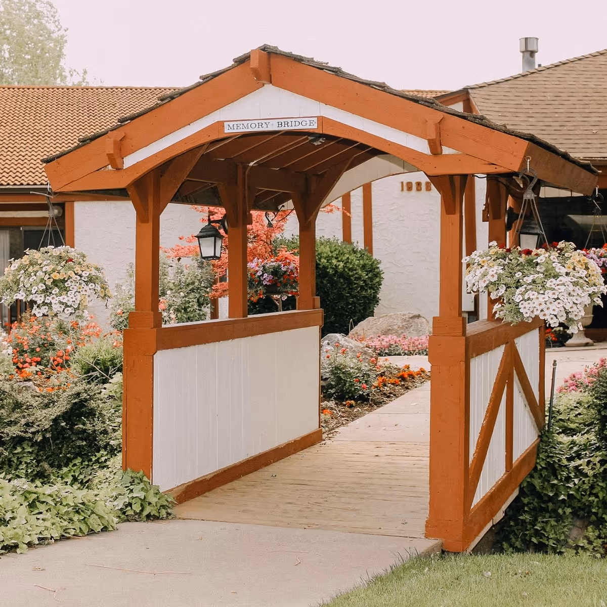 Covered wooden garden bridge labeled "Memory Bridge" with hanging flower baskets and landscaping leading toward a building.