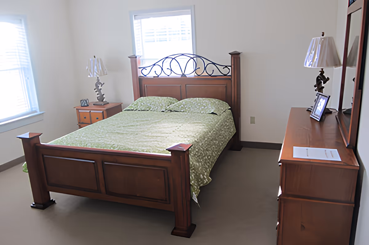 A bedroom with a wooden bed frame featuring a decorative metal headboard, a green floral bedspread, two pillows, a wooden nightstand with a lamp and a small picture frame, and a wooden dresser with a lamp, a picture frame, and a mirror. The room has two windows with white blinds and light-colored walls.