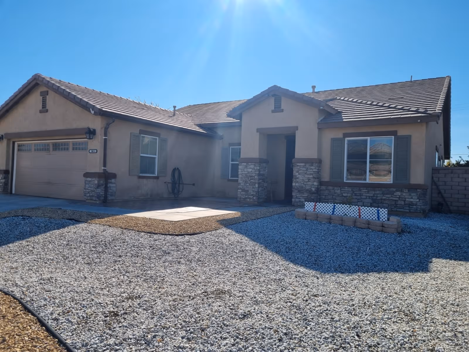 Single-story house with a beige exterior, stone accents, and a tiled roof under a clear blue sky. The front yard is covered with gravel and has a small garden bed bordered with bricks. There is a two-car garage on the left side and a covered entrance in the center.