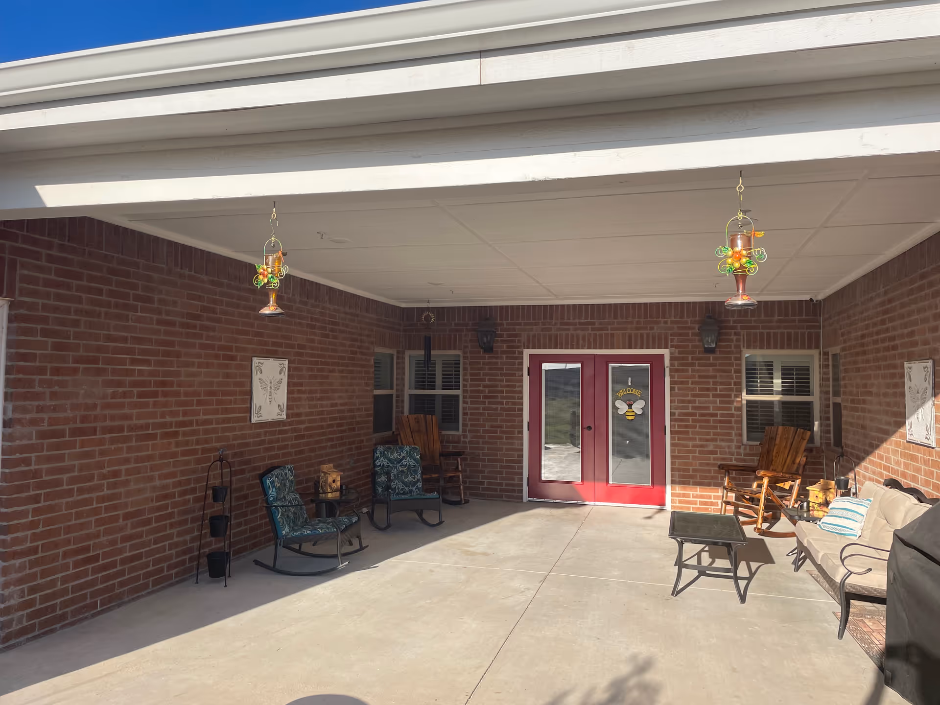 Covered brick patio with seating, hanging decorative lanterns, and double red doors with a bee decoration.