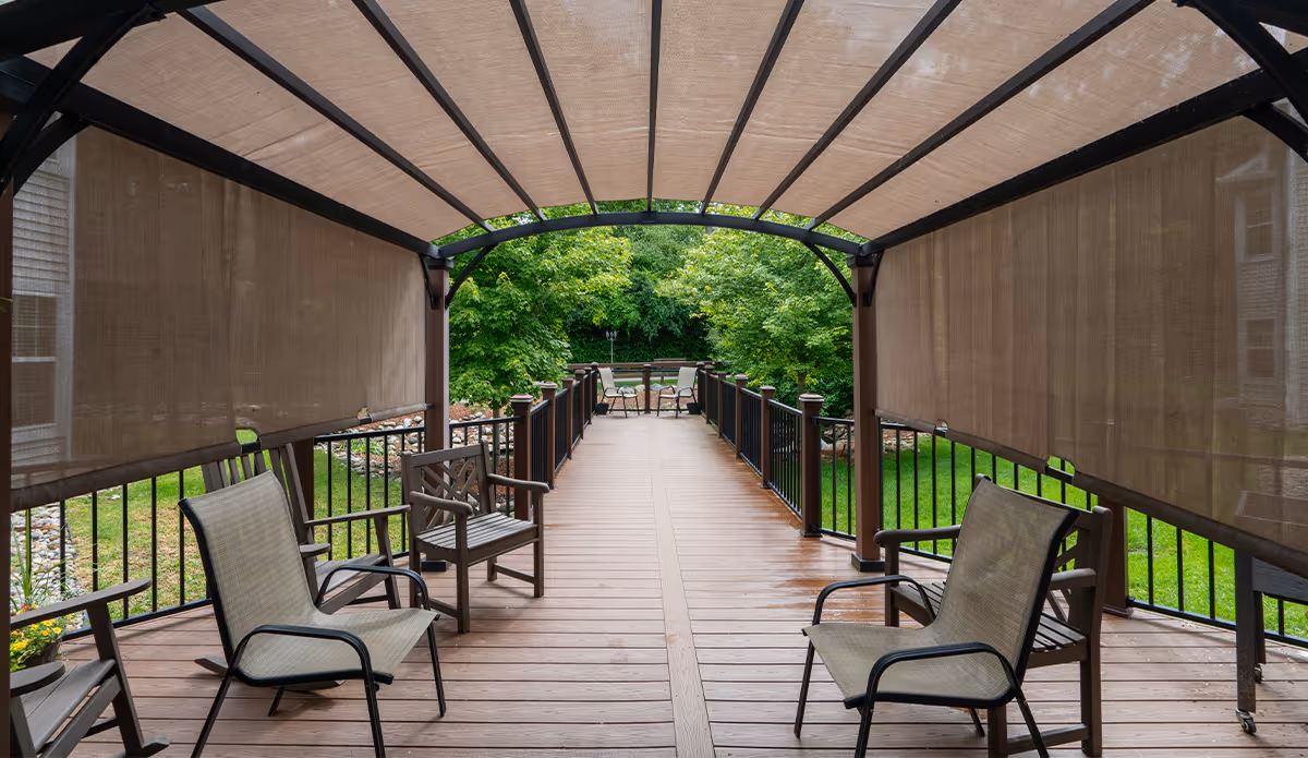Covered outdoor wooden deck with several chairs arranged along the sides and at the far end, surrounded by green grass and trees.