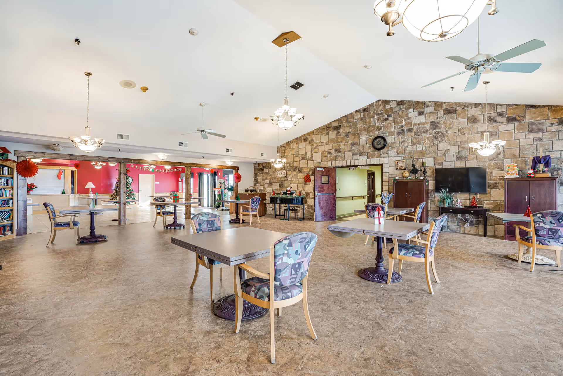 Spacious senior living communal dining and activity room with tables and chairs, a stone accent wall, TV and holiday decorations.