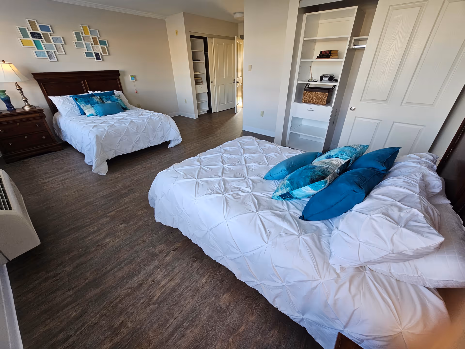 A bright bedroom with two beds, each made with white comforters and blue decorative pillows. There is a wooden nightstand with a lamp between the beds. The room has wood flooring, a white built-in shelving unit, and a closet with open doors. Colorful abstract wall art is displayed above one of the beds.