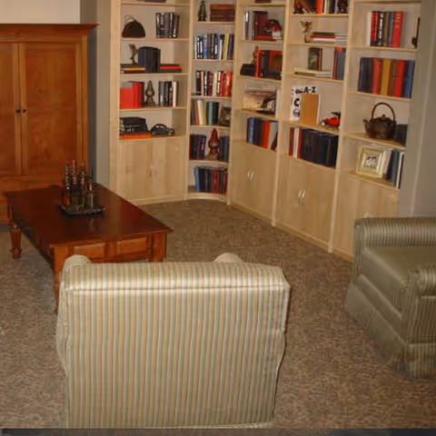 A cozy living room area with two striped armchairs facing a wooden coffee table. Behind the table is a wooden cabinet, and the walls are lined with built-in bookshelves filled with books and decorative items.