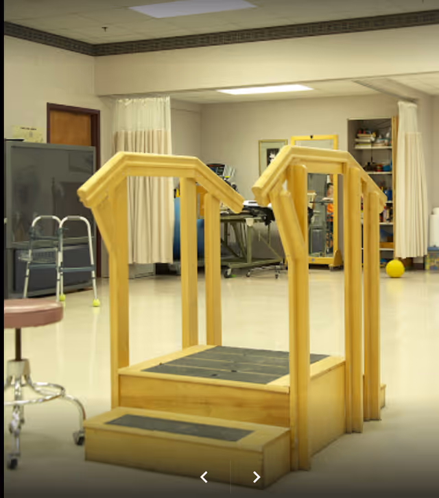 Indoor physical therapy room with wooden parallel bars for walking exercises, a walker, a stool, and various therapy equipment in the background.