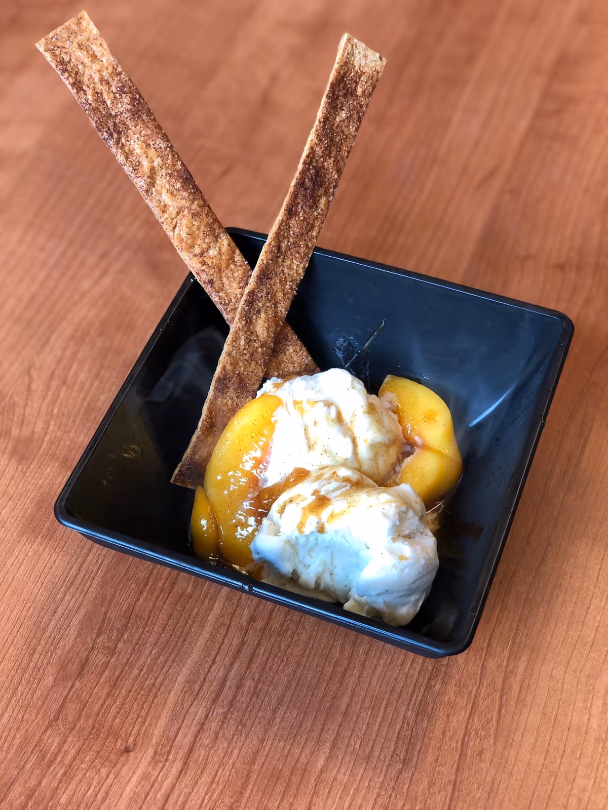 A black square bowl containing two scoops of vanilla ice cream topped with peach slices and syrup, garnished with two long cinnamon sugar sticks, placed on a wooden surface.