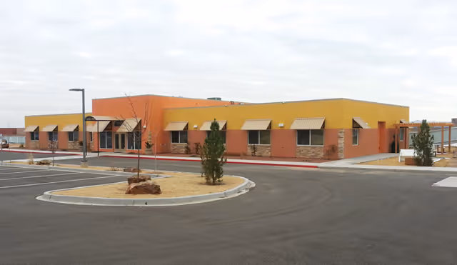 Exterior view of a single-story senior living facility building with a yellow and orange facade, multiple windows with awnings, a parking lot in front, and some small trees and landscaping.