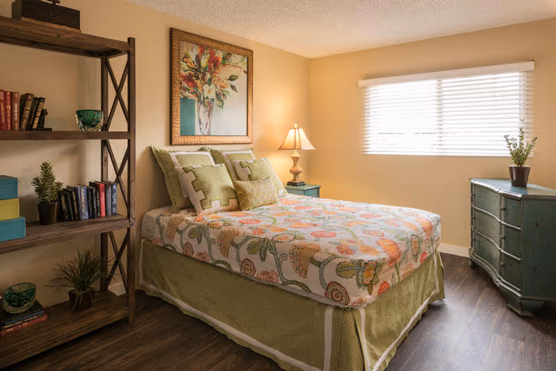 A cozy bedroom with a bed covered in a floral patterned bedspread and green pillows. There is a wooden bookshelf with books and decorative items on the left side, a framed floral painting above the bed, a bedside table with a lamp, and a green dresser with a potted plant near a window with white blinds.
