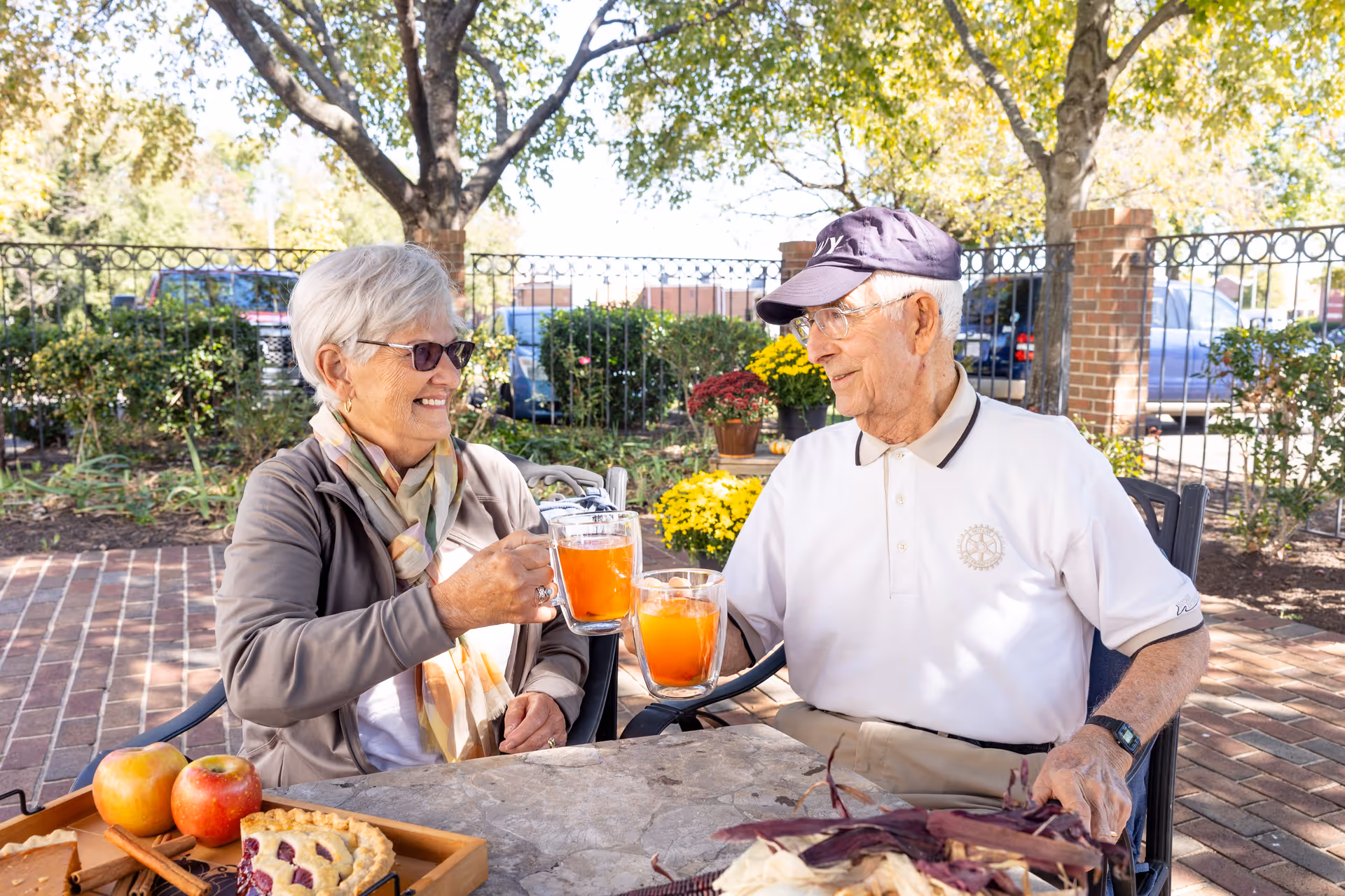 An elderly woman and man sitting outdoors at a table, smiling and clinking glasses filled with an orange beverage. The table has a tray with apples and a slice of pie. They are surrounded by trees, flowers, and a metal fence in a sunny garden setting.